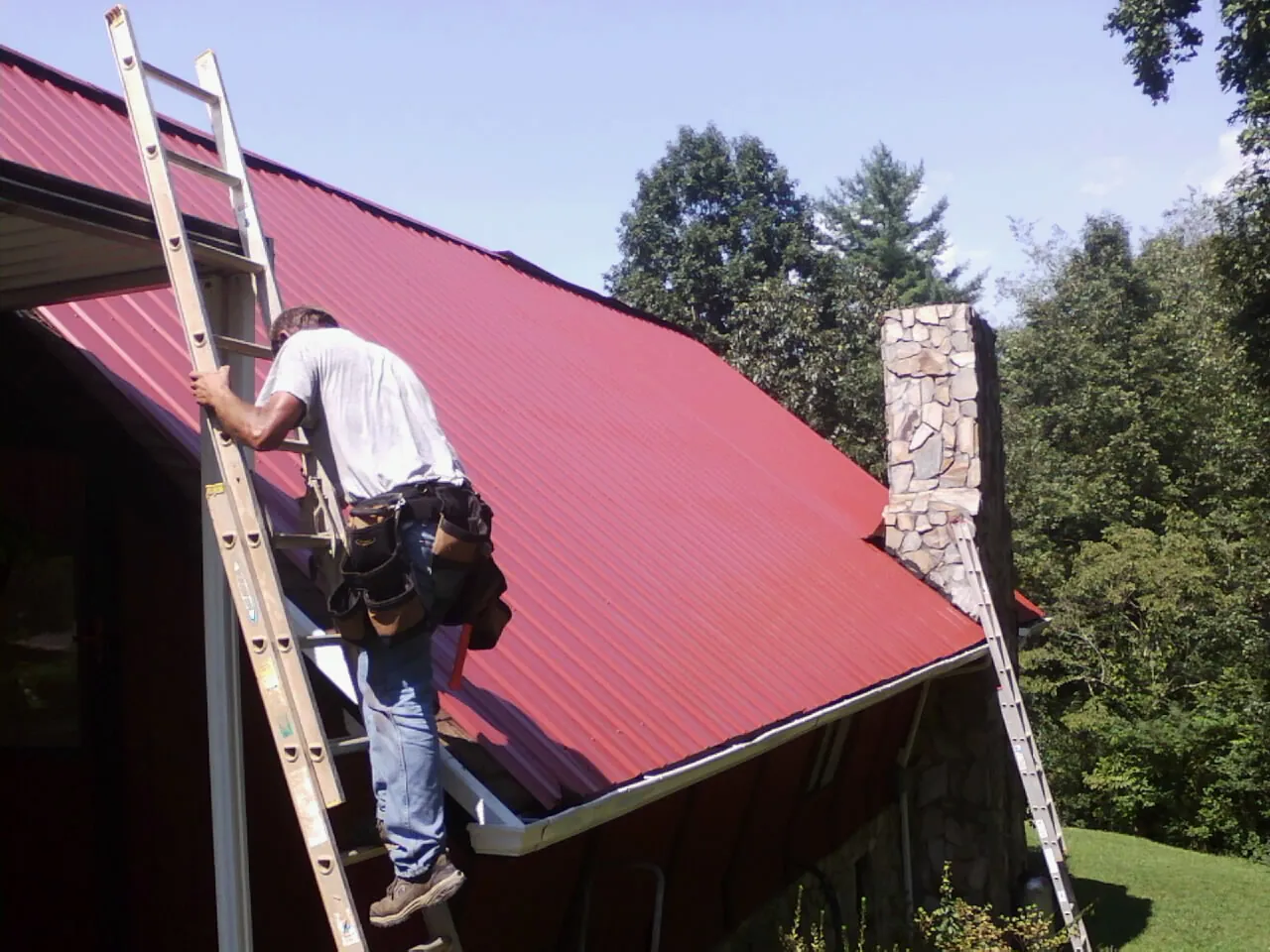 Roofer on a ladder beside a red metal roof and stone chimney — Soffit & Fascia Repair by Roofing Brothers in Braxton