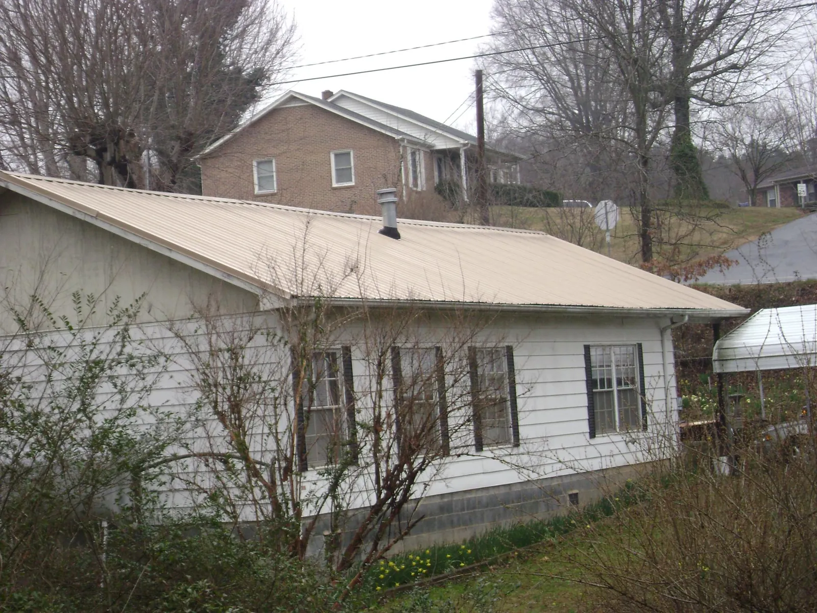 Tan metal roof on a white sided house set into a hillside — Commercial Roofing by Roofing Brothers in Braxton